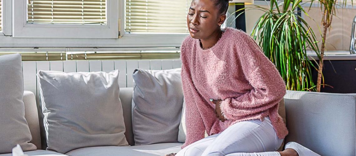 Young black woman suffering from stomachache on sofa at home. Woman sitting on bed and having stomach ache. Young woman suffering from abdominal pain while sitting on sofa at home