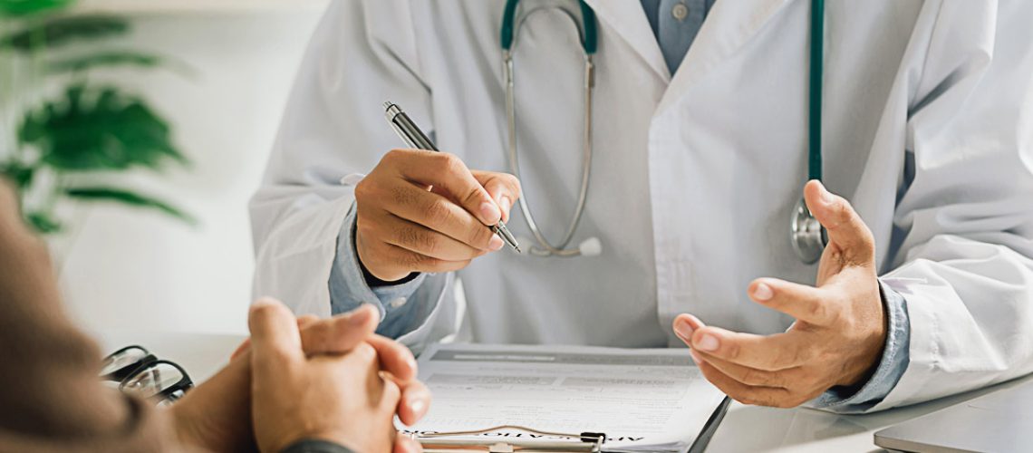 A professional physician in a white medical uniform talks to discuss results or symptoms and gives a recommendation to a male patient and signs a medical paper at an appointment visit in the clinic.